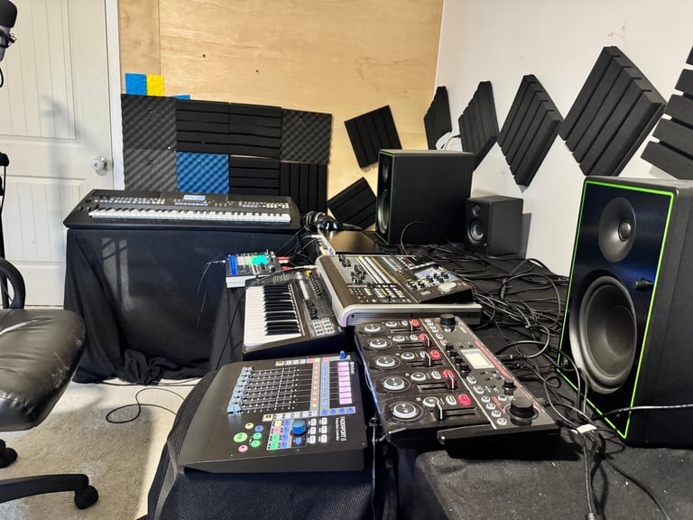 Professional music production studio setup with keyboards, mixing console, audio interfaces, studio monitors, and acoustic panels against wooden and white walls
