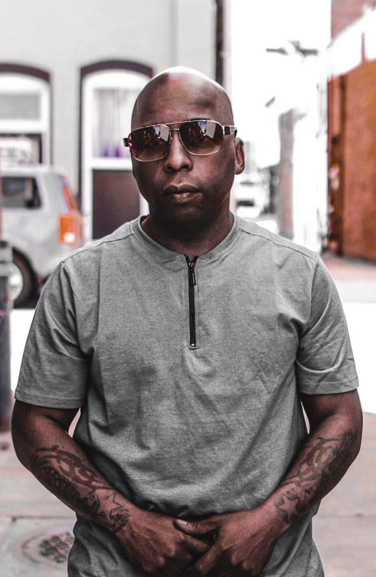 Man wearing gray henley shirt and sunglasses standing outdoors with arms crossed, parked cars visible in background
