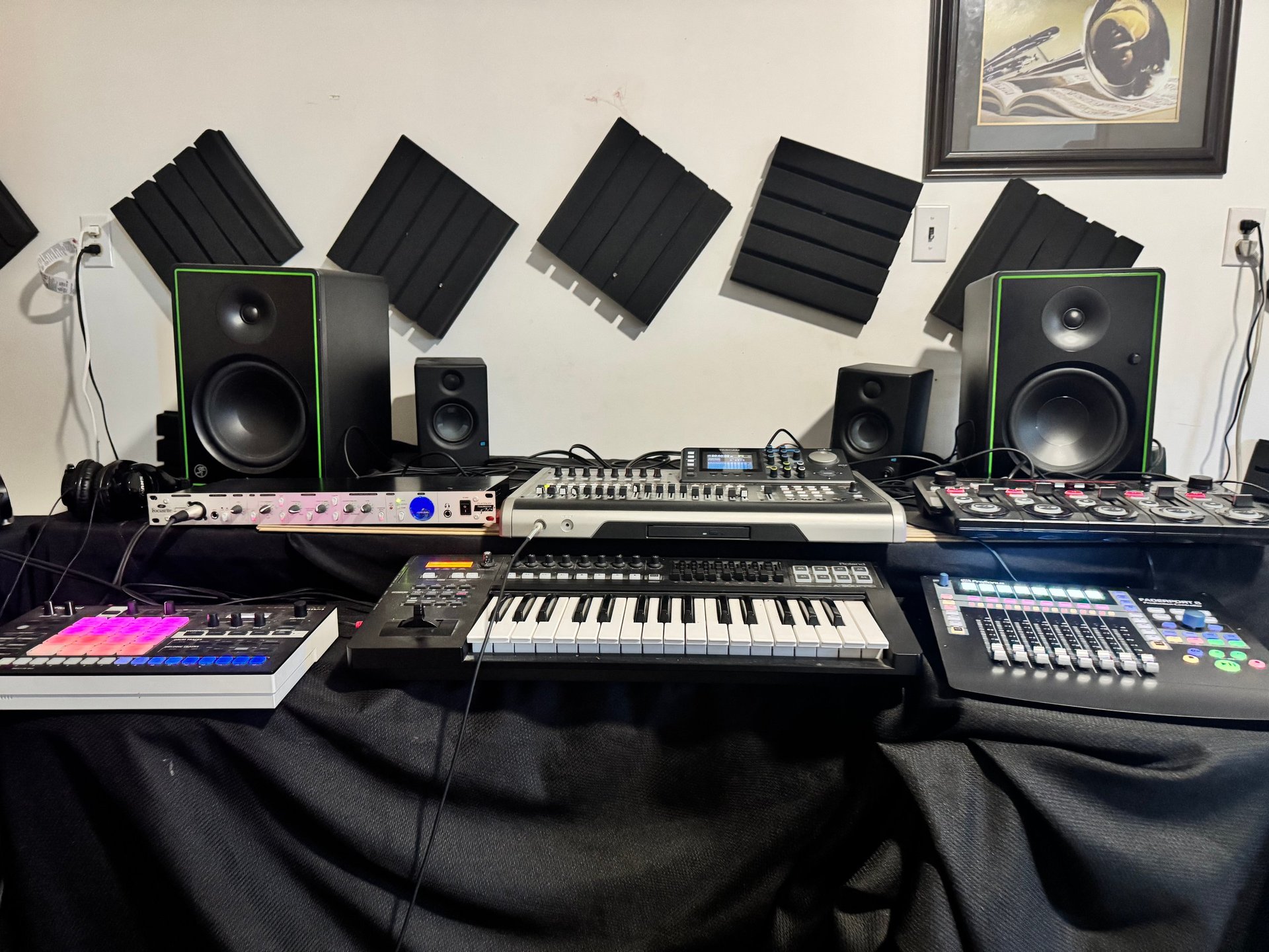 Professional music production studio setup with keyboards, synthesizers, audio monitors, and acoustic panels on a wall