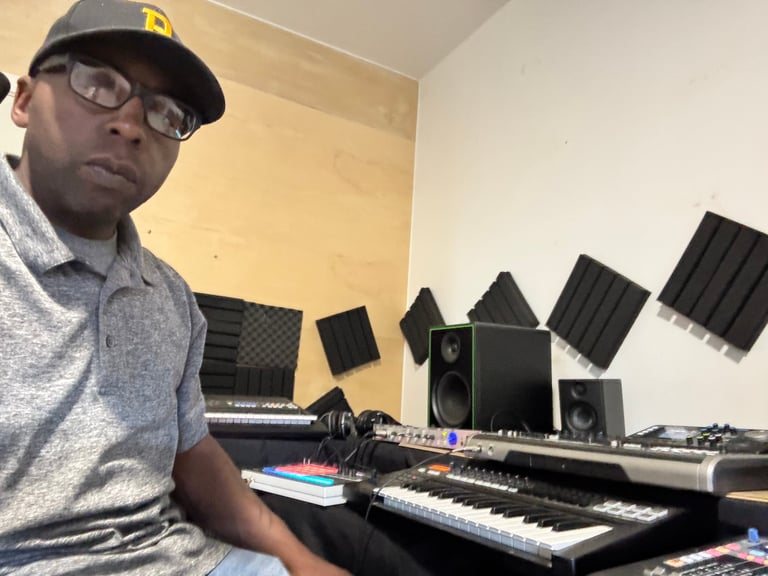 Person wearing glasses and cap seated at music production setup with keyboards and studio monitors on wall