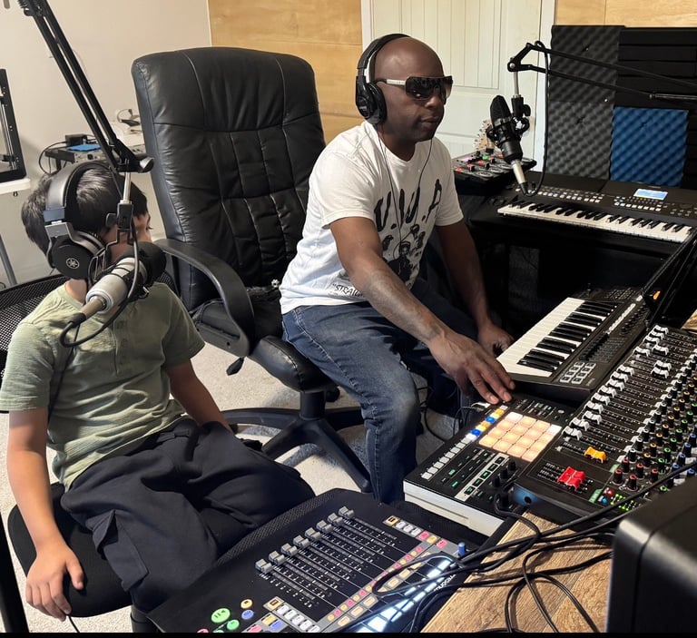 Man with headphones and sunglasses at music production setup with keyboard and mixing equipment, teaching a young student